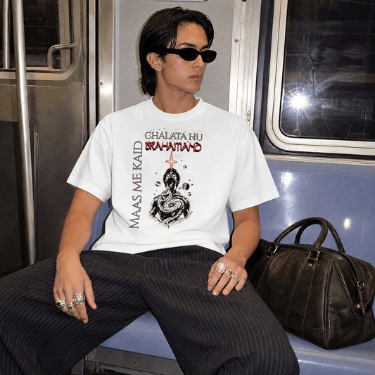 A person sitting on a subway train wearing a white graphic T-shirt with black and red text and artwork, paired with dark pinstriped pants and a dark brown bag beside them.