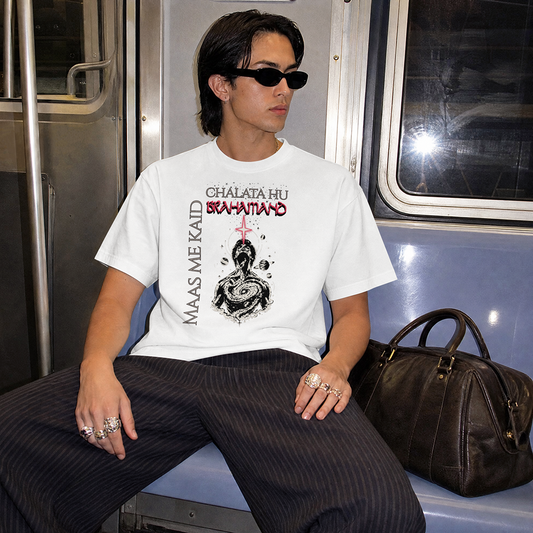 A person wearing a white graphic T-shirt with black and red text and artwork, seated on a train. The shirt reads  'MAAS ME KAID CHALATA HU BRAHAMAND'