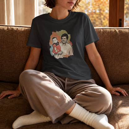 A person sitting on a brown couch wearing a dark charcoal graphic t-shirt and beige sweatpants. The t-shirt features a colorful graphic print of a man and child.